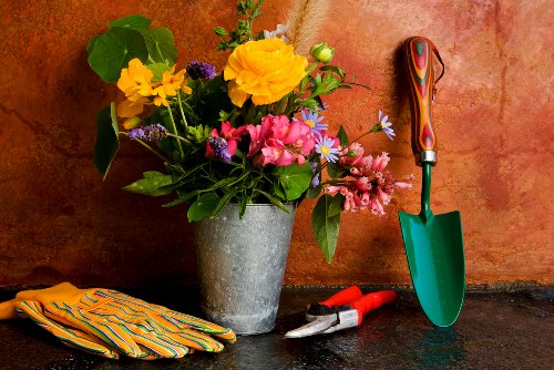 A close-up view of a garden workspace showing a variety of gardening tools and materials placed on a dark, textured surface. In the center, there is a large, round, grey concrete flower pot filled with an assortment of freshly cut flowers, including yellow, pink, purple, and white blossoms, along with green foliage. To the right of the pot, a small green-handled trowel with a shiny metal blade stands upright, and next to it are red-handled pruning shears resting on the surface. On the left side, a pair of striped garden gloves are neatly folded. The background features a warm, reddish-brown wall with a subtle textured finish, creating a professional and natural gardening scene that subtly reflects outdoor planting and maintenance activities typical of Raynes Park in the London borough of Merton. This setup suggests gardening services such as plant care, flower arranging, and outdoor maintenance are provided by Lawn Mowing Raynes Park, aligning with the landscaping and garden upkeep theme.